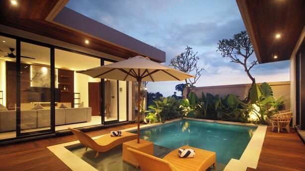 A modern indoor pool area with lounge chairs and an umbrella, surrounded by wooden flooring and glass walls.