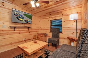 Ein Ferienhausinnenraum mit an der Wand montiertem Fernseher, Deckenventilator, Holztisch, Stuhl und einem Fenster mit Jalousien.