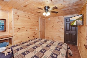 Ein Zimmer mit Holzwänden, einem Deckenventilator, einem Bett mit gemustertem Überwurf und einem Fernseher an der Wand sowie einer Tür.
