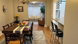 A dining area with a wooden table, chairs, and a chandelier. There is a view of a living room with a sofa and a plant.