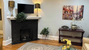 A living room with a fireplace, a flat-screen TV mounted above it, a painting of a street scene, and a small table with a vase of flowers.