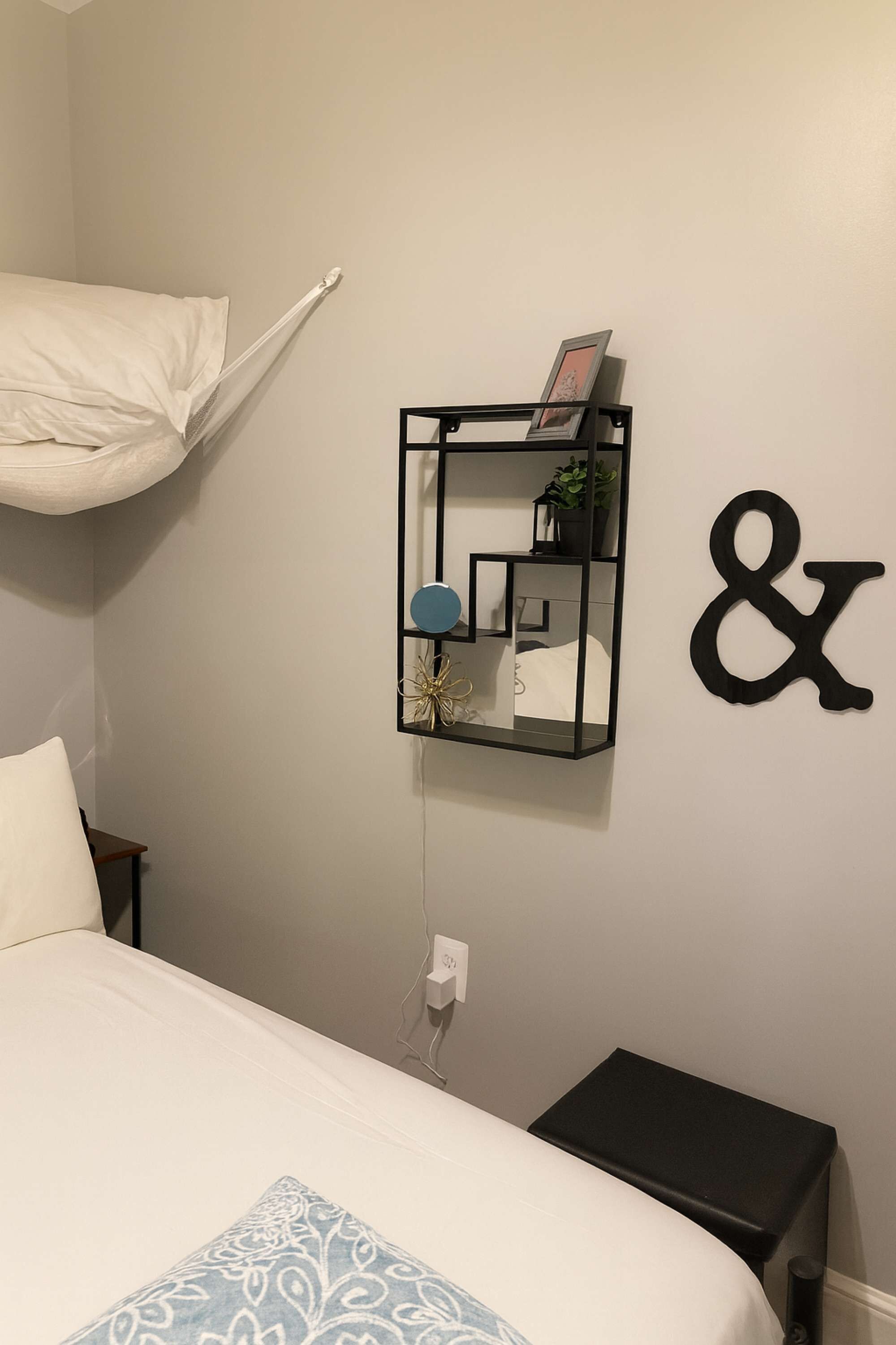 A bedroom with a bed, a bedside table, a wall-mounted shelf with decorative items, and a wall decoration.