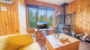 Ein gemütliches Blockhaus-Interieur mit Holzausstattung, einem Steinofen, einem Fernseher und einem großen Fenster mit Blick auf Bäume.