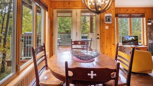 Ein Holzinterieur mit Esstisch, Stühlen, einer Deckenleuchte und großen Fenstern mit Blick auf Bäume.