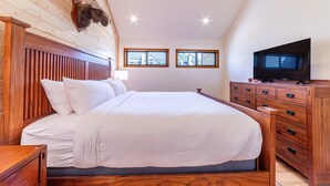 A bedroom with a wooden headboard, a large bed with white bedding, a wooden dresser with a flat-screen TV, and two windows with a view of buildings.