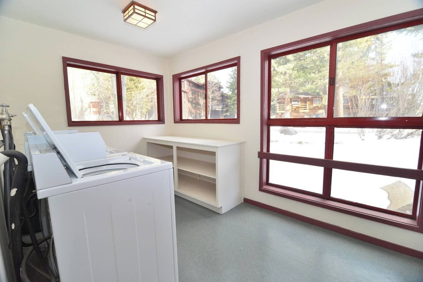 A laundry room with a washing machine, a drying rack, and a white shelf unit. There are three windows with wooden frames, and the room has a concrete floor.