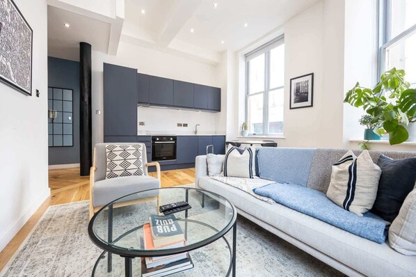 Un salón moderno con una mesa de centro de vidrio, un sofá con cojines estampados y vistas a una cocina al fondo.
