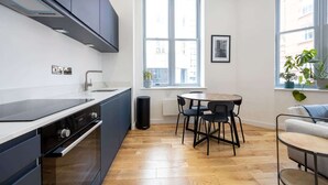 A modern kitchen with dark cabinets, a white countertop, and a built-in oven. There is a window with a view of buildings outside, a round table with chairs, and a potted plant on the windowsill. - Azure Suite 1BR (Greater Manchester)