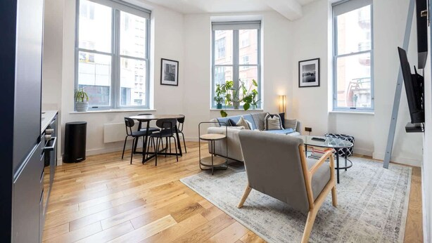 A modern living room with a sofa, coffee table, and dining area. There are large windows providing natural light and views of the city. - Azure Suite 1BR (Greater Manchester)
