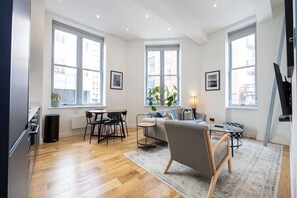 Una sala de estar moderna con sofá, mesa de centro y comedor. Amplias ventanas ofrecen luz natural y vistas de la ciudad.