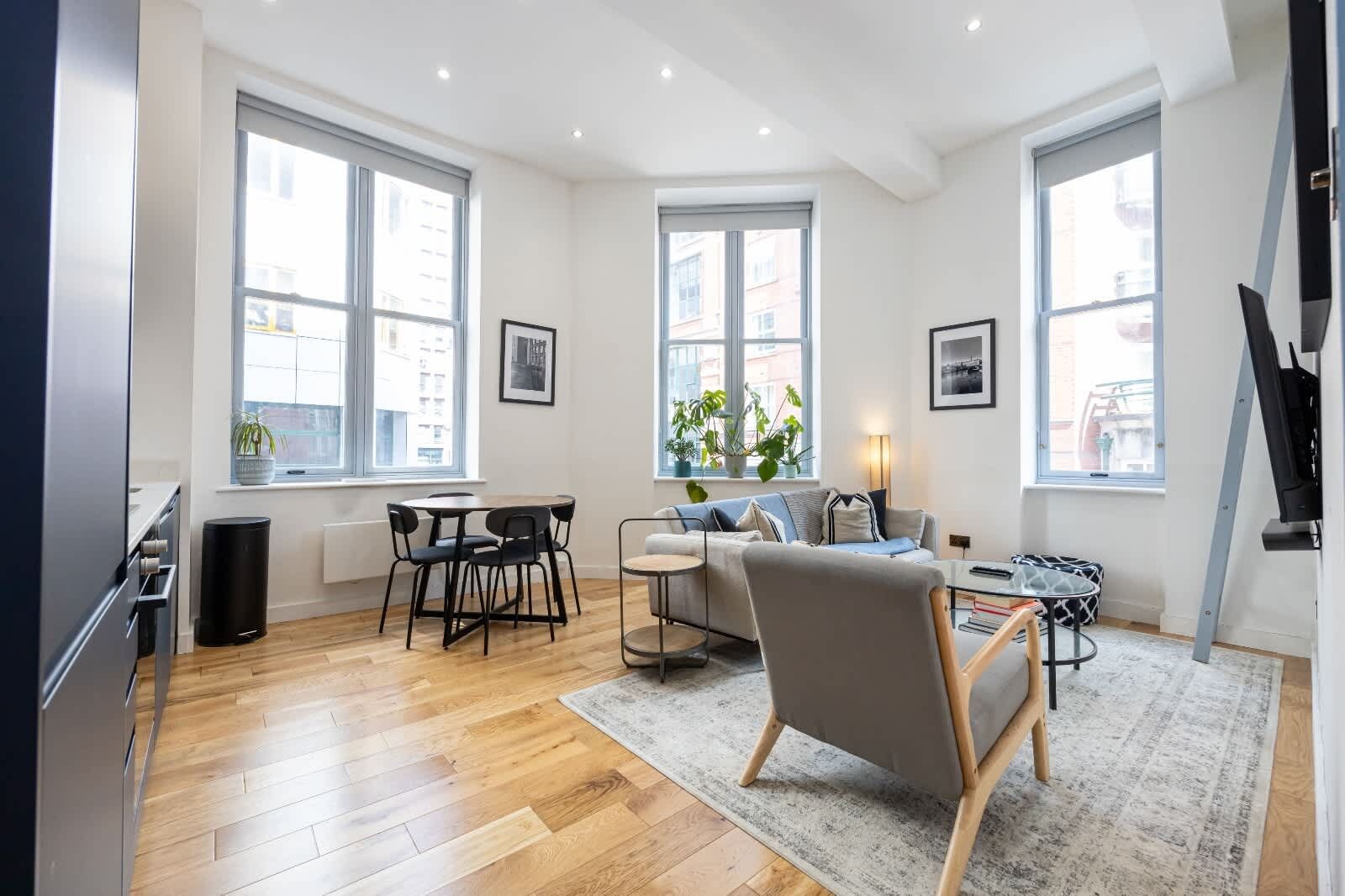 Una sala de estar moderna con sofá, mesa de centro y comedor. Amplias ventanas ofrecen luz natural y vistas de la ciudad.
