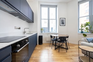 Une cuisine moderne avec des meubles de couleur sombre, un plan de travail blanc et un four encastré. Une fenêtre offre une vue sur les bâtiments environnants, et on y trouve une table ronde avec des chaises, ainsi qu’une plante en pot posée sur le rebord de la fenêtre.
