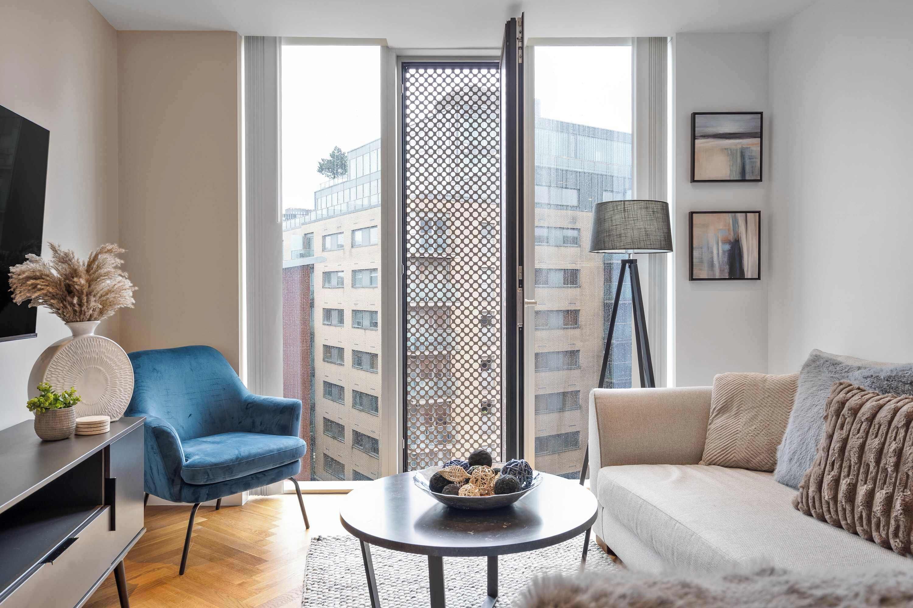 Een moderne woonkamer met een blauwe fauteuil, een ronde salontafel met decoratieve objecten, een beige bank met een plaid, en een vloerlamp.