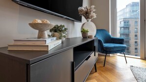 Ein modernes Wohnzimmer mit einem großen Flachbildfernseher an der Wand, einem dunklen Holzkonsole und dekorativen Gegenständen, einem blauen Sessel und Blick auf eine Stadtlandschaft durch große Fenster.