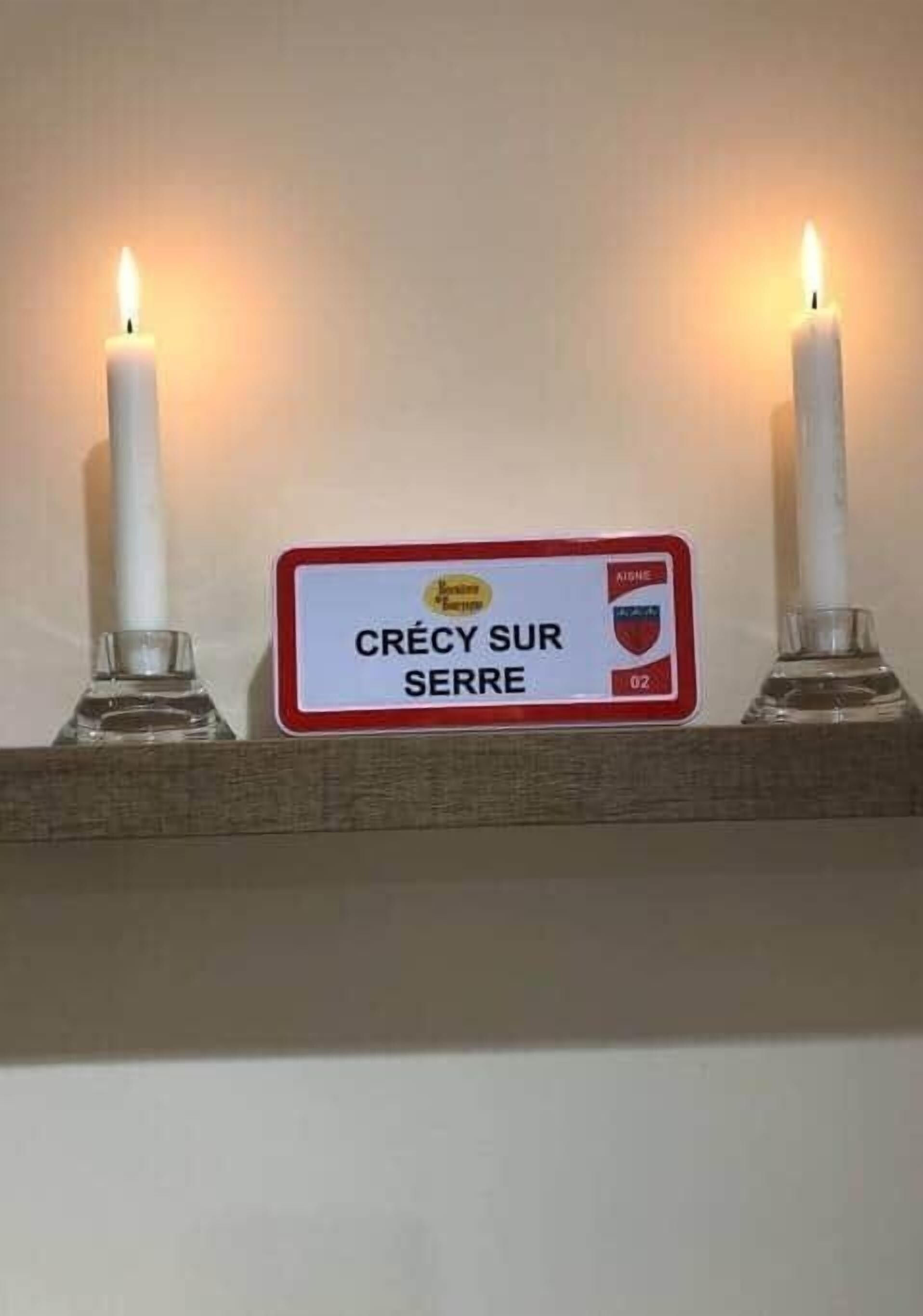 A sign with the text "CRÉCY SUR SERRE" and a number "02" on a shelf with two lit candles.