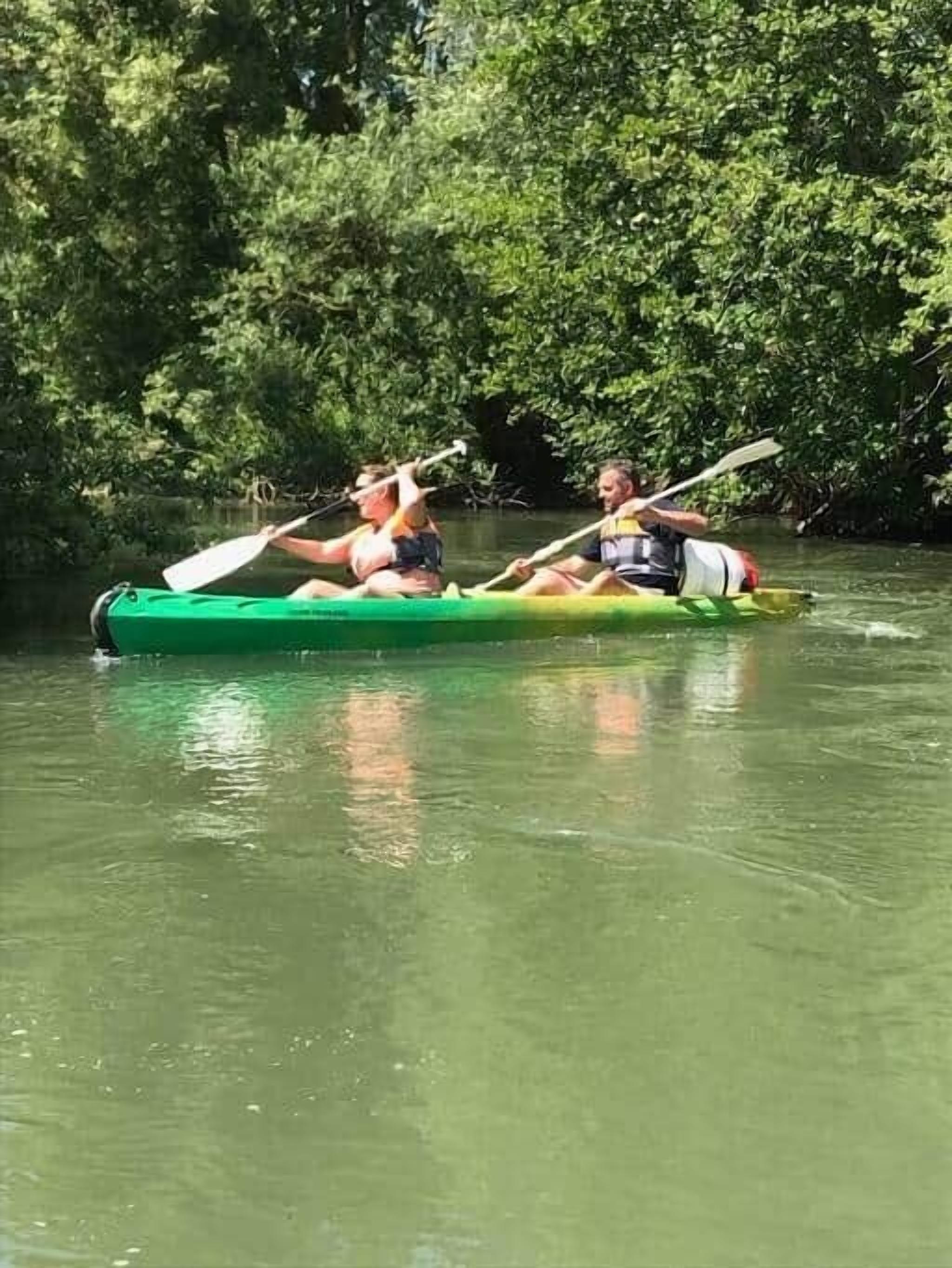 Deux personnes pratiquent le kayak dans un canoë vert sur une rivière paisible, entourée d’une végétation luxuriante.