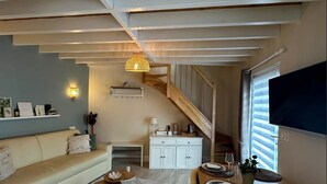 Ein großzügiger Wohnbereich mit hohen Decken, einer Holztreppe, einem Sofa, einem Esstisch mit Stühlen und einem an der Wand montierten Fernseher.