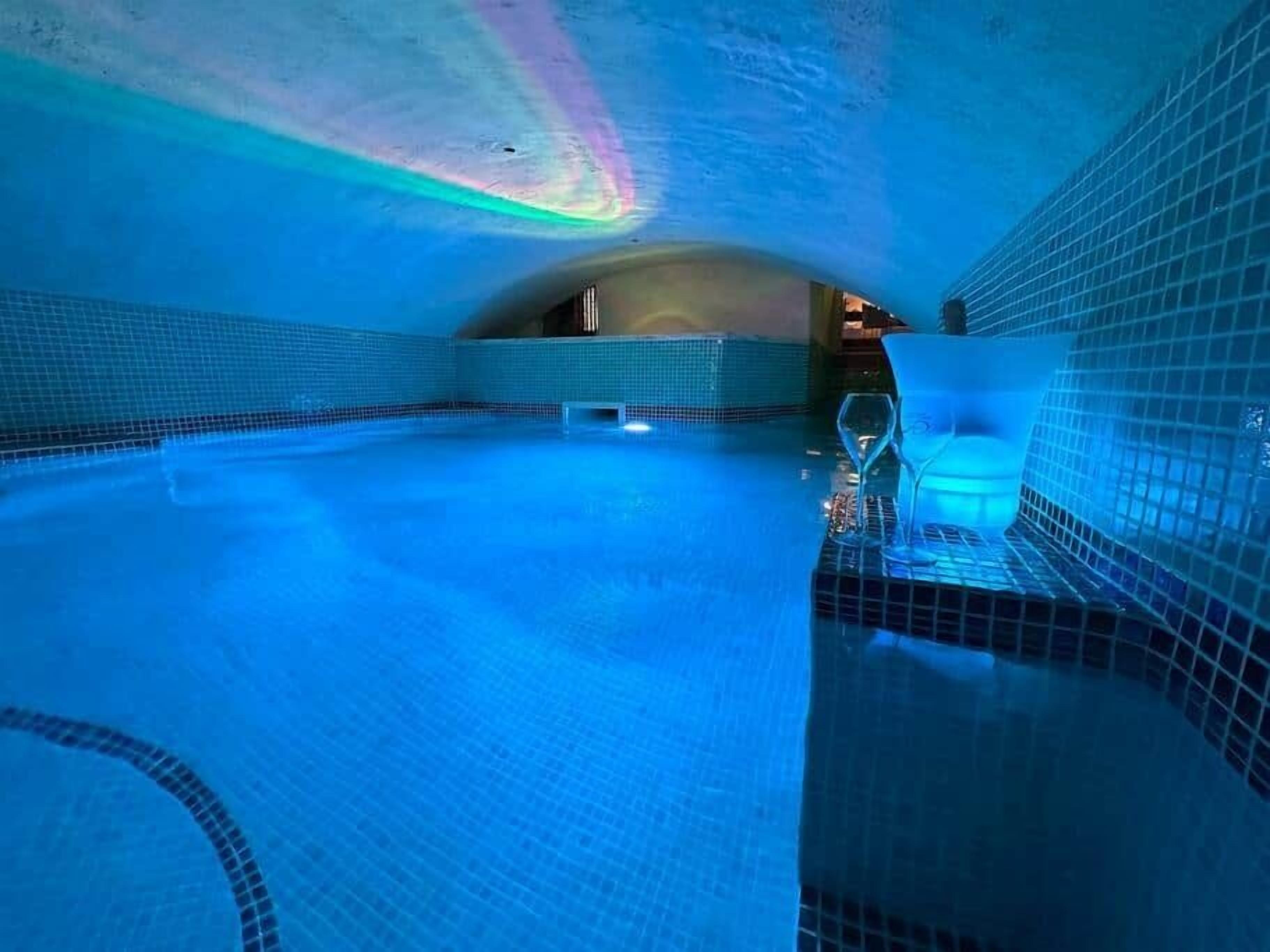 Une piscine intérieure avec un sol et des murs carrelés, une cloison en verre et un plafond doté d’un luminaire.
