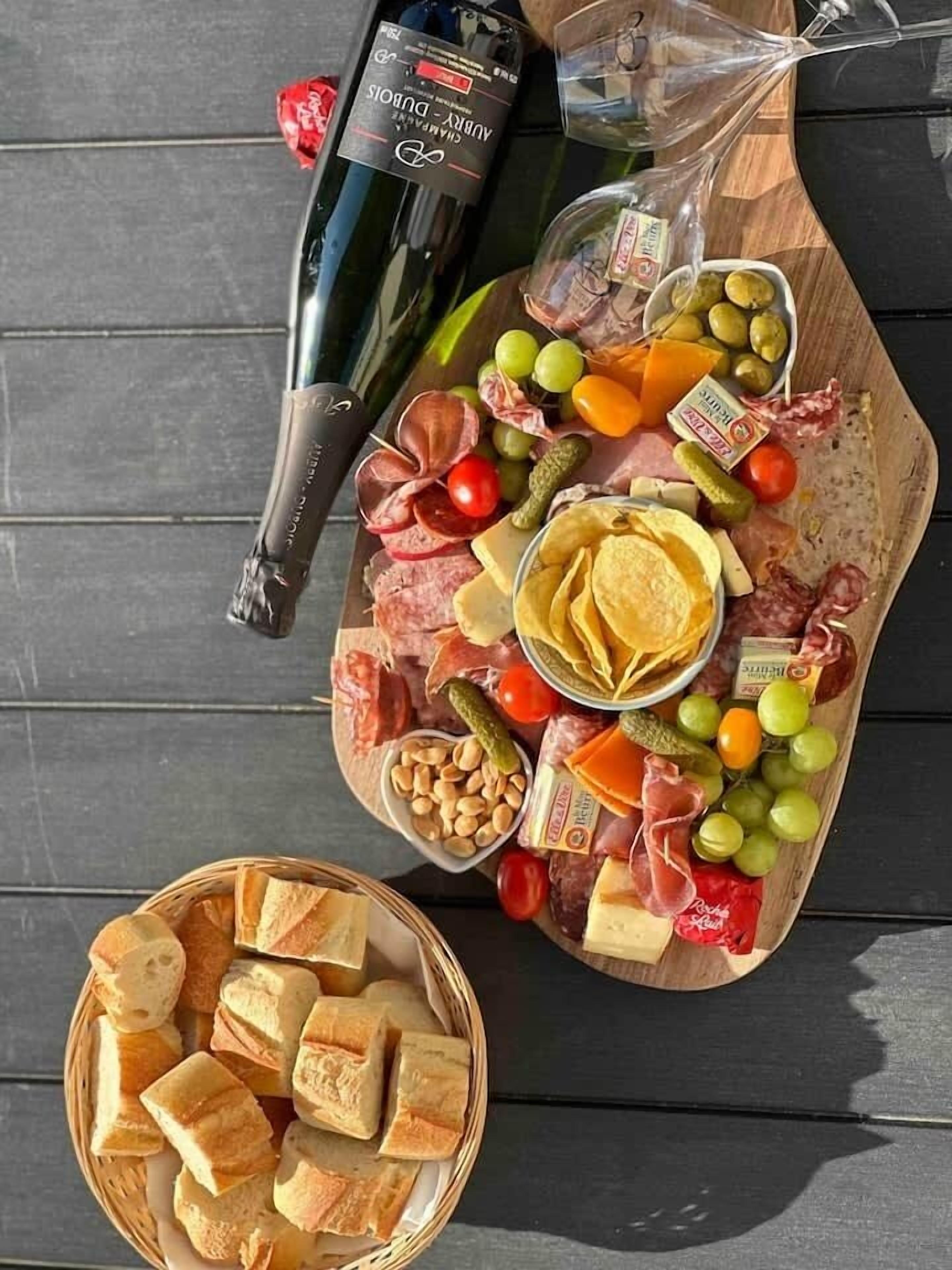 Une planche de charcuterie garnie de différentes sortes de viandes, de fromages et de fruits, accompagnée d’une bouteille de vin et d’un panier de pain.