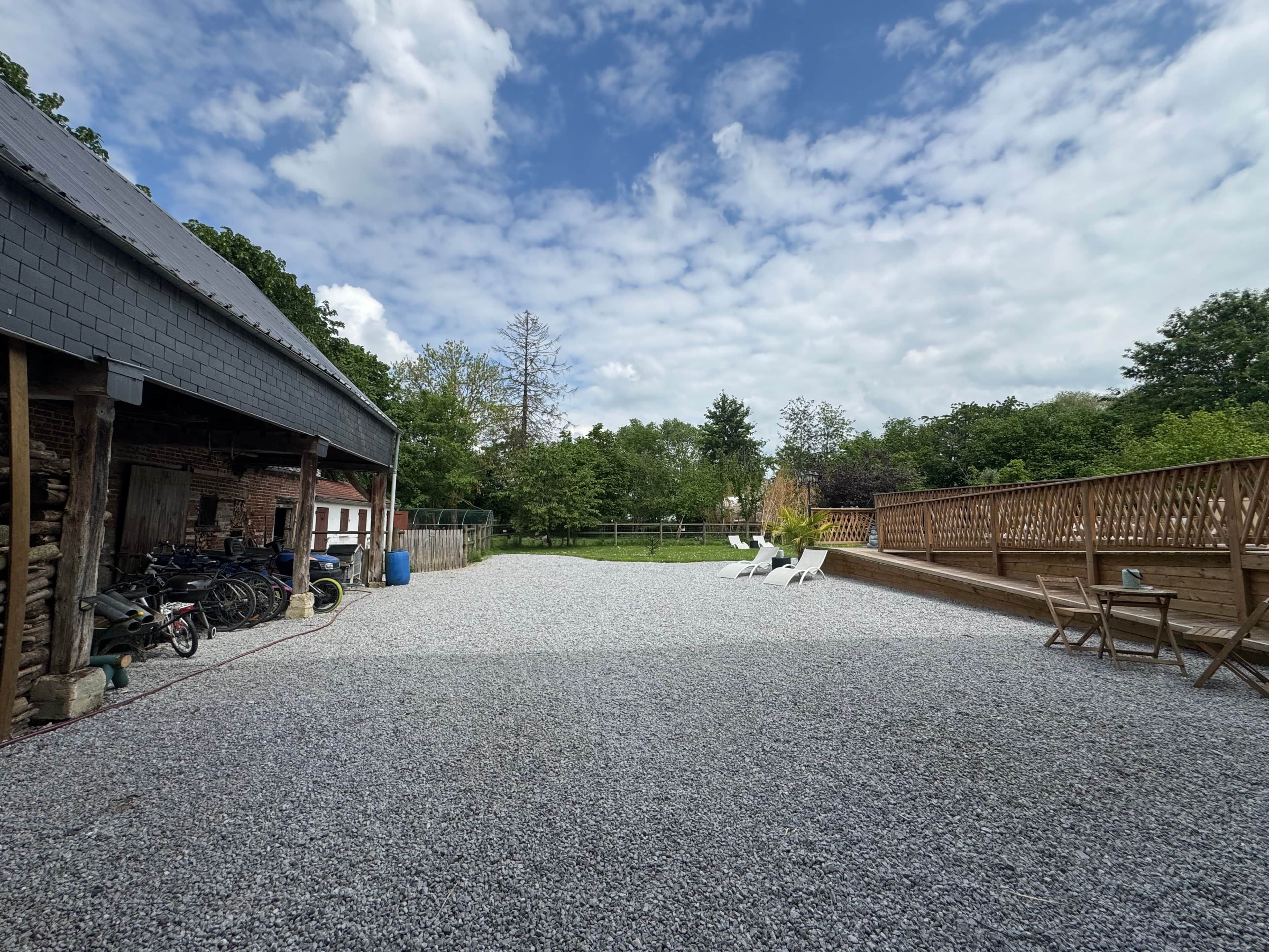 A gravel courtyard with a wooden fence, a covered area with bicycles, and a view of trees and sky.