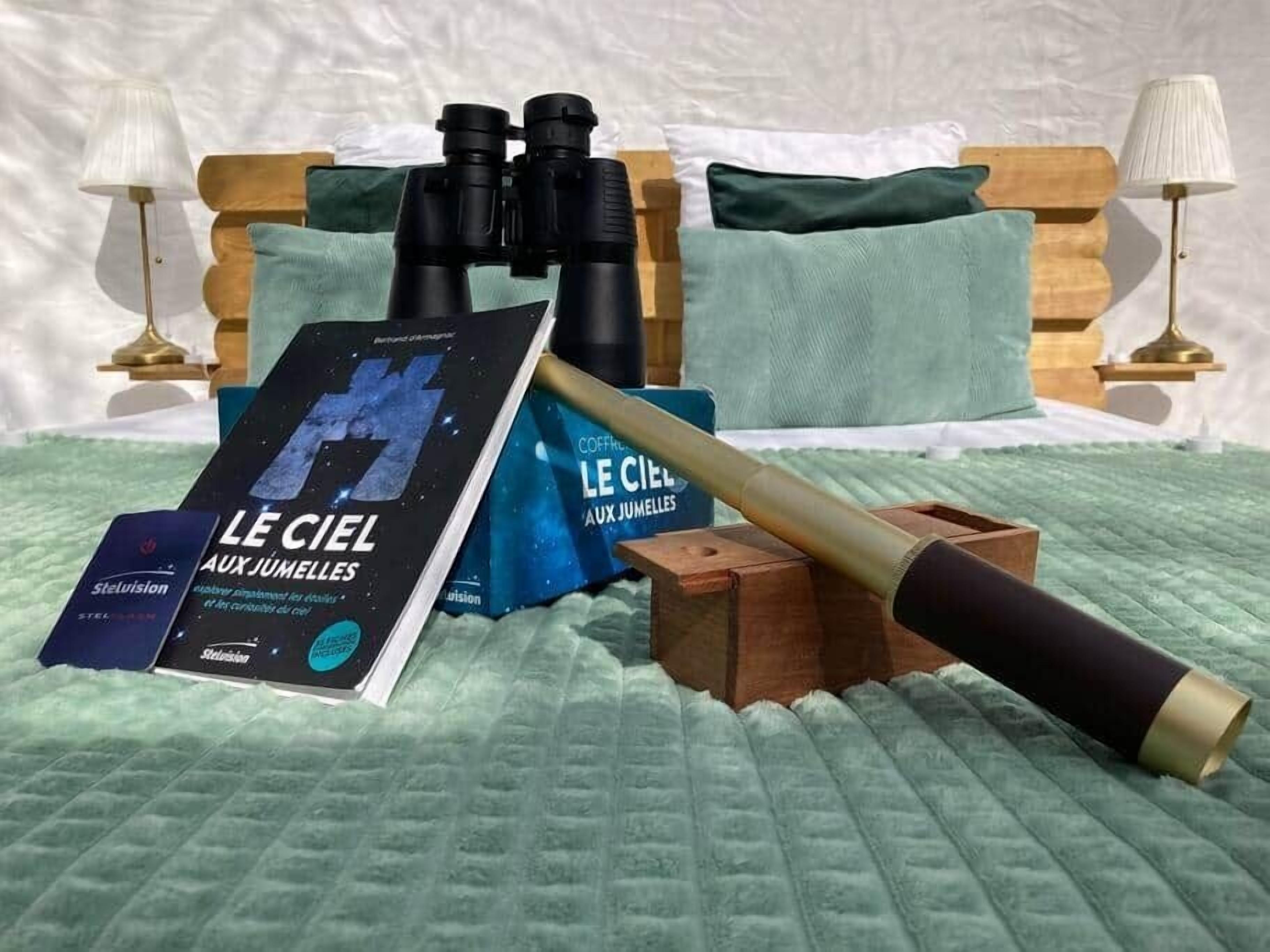 Een bed met een groene sprei, een verrekijker, een boek getiteld 'LE CIEL AUX JUMELLES' en een houten doos met een gouden voorwerp.