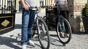 Ein Mann und eine Frau stehen mit Fahrrädern vor einem Tor mit einer Ziegelwand und einer Tafel.