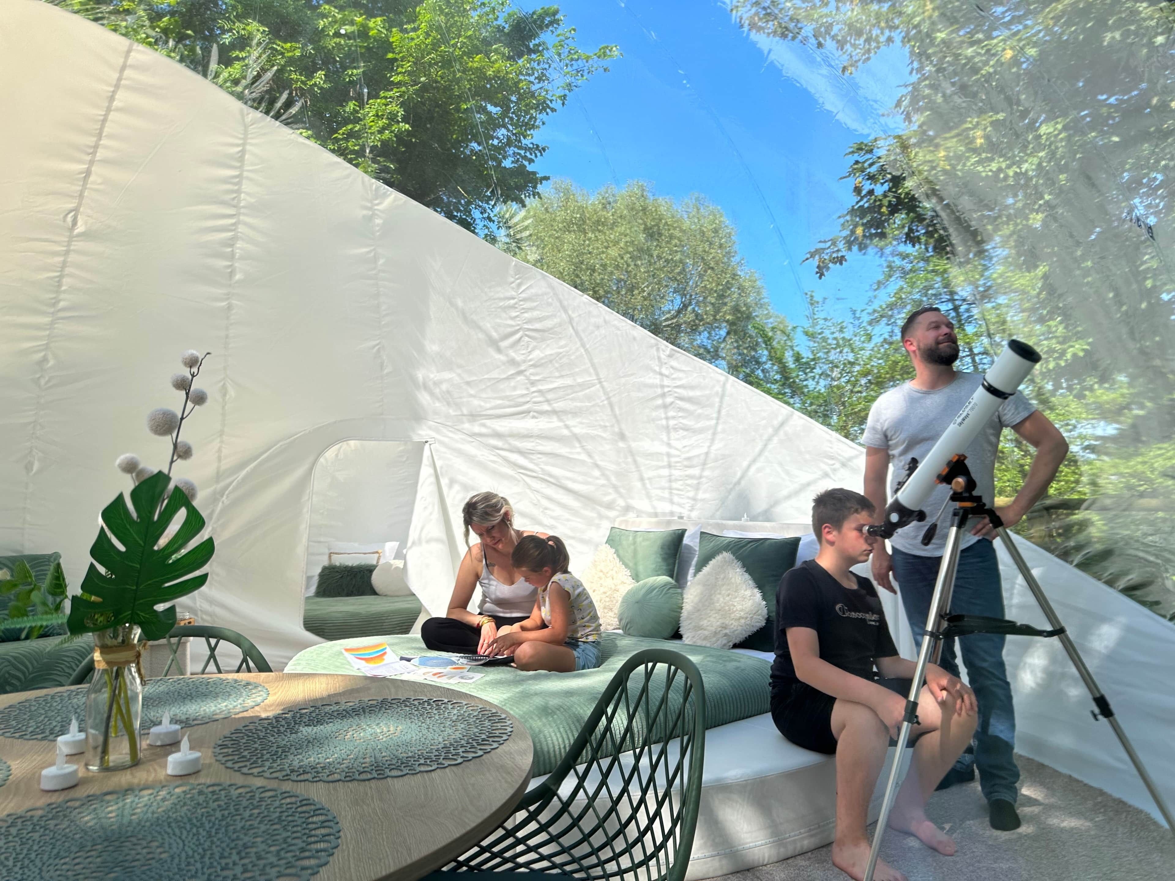 Une tente dotée d’un espace salon avec des coussins verts, d’un télescope et d’une table ornée de bougies et de papiers.