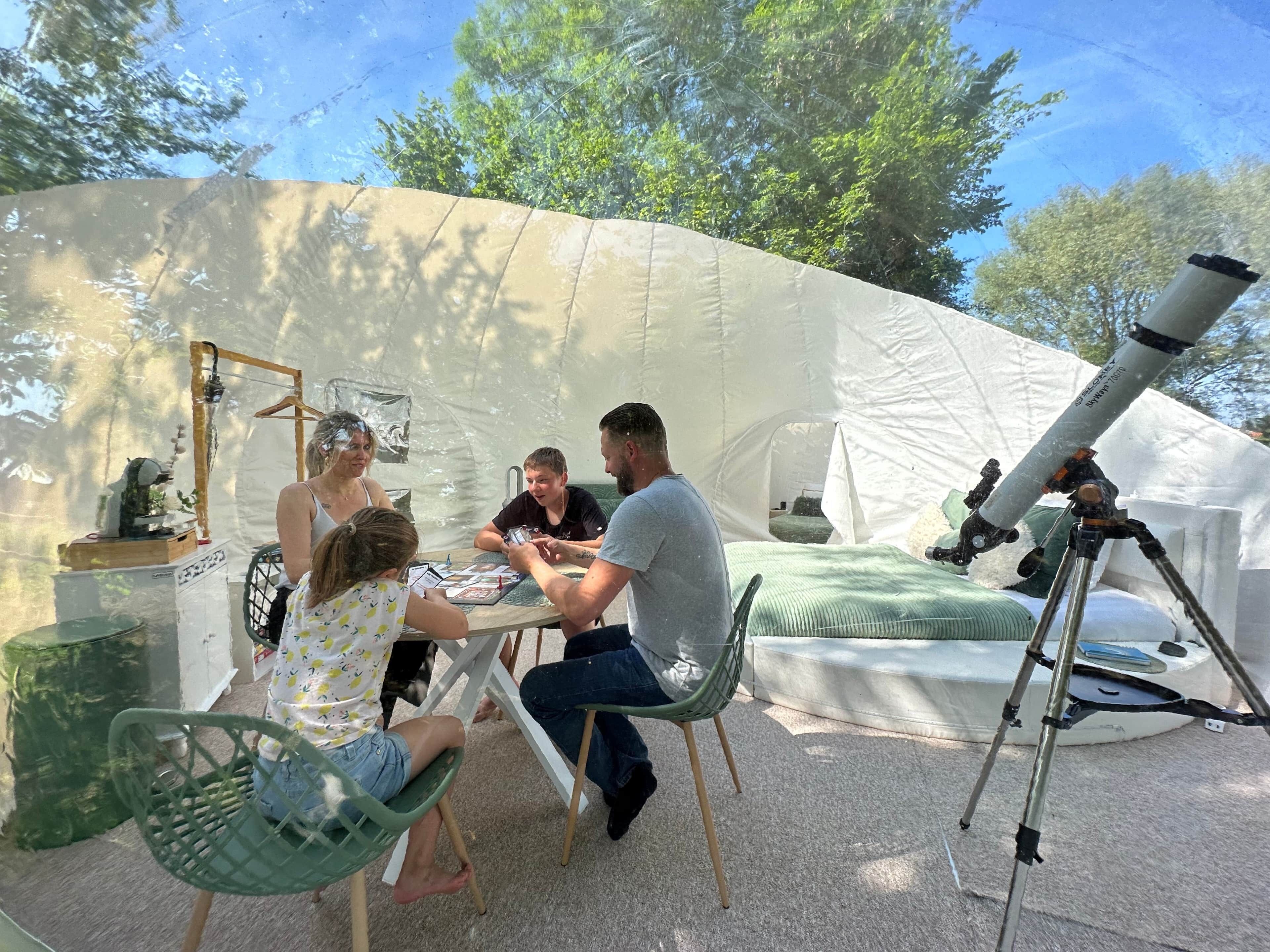 A group of people seated at an outdoor table with a telescope and a tent in the background.