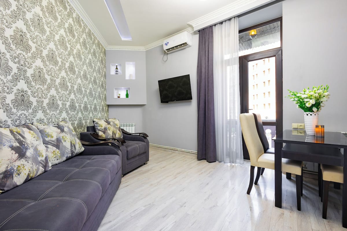 Ein Wohnzimmer mit grauem Sofa, einem Esstisch mit Stühlen, einem an der Wand befestigten Flachbildfernseher und einem Fenster mit Vorhängen.
