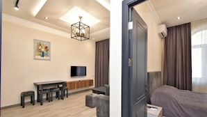 A modern hotel room with a large bed, a desk with a flat-screen TV, a dining area with stools, and a painting on the wall.