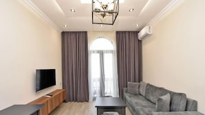 A modern living room with a flat-screen TV, a grey sofa, a small table, and a large window with curtains.