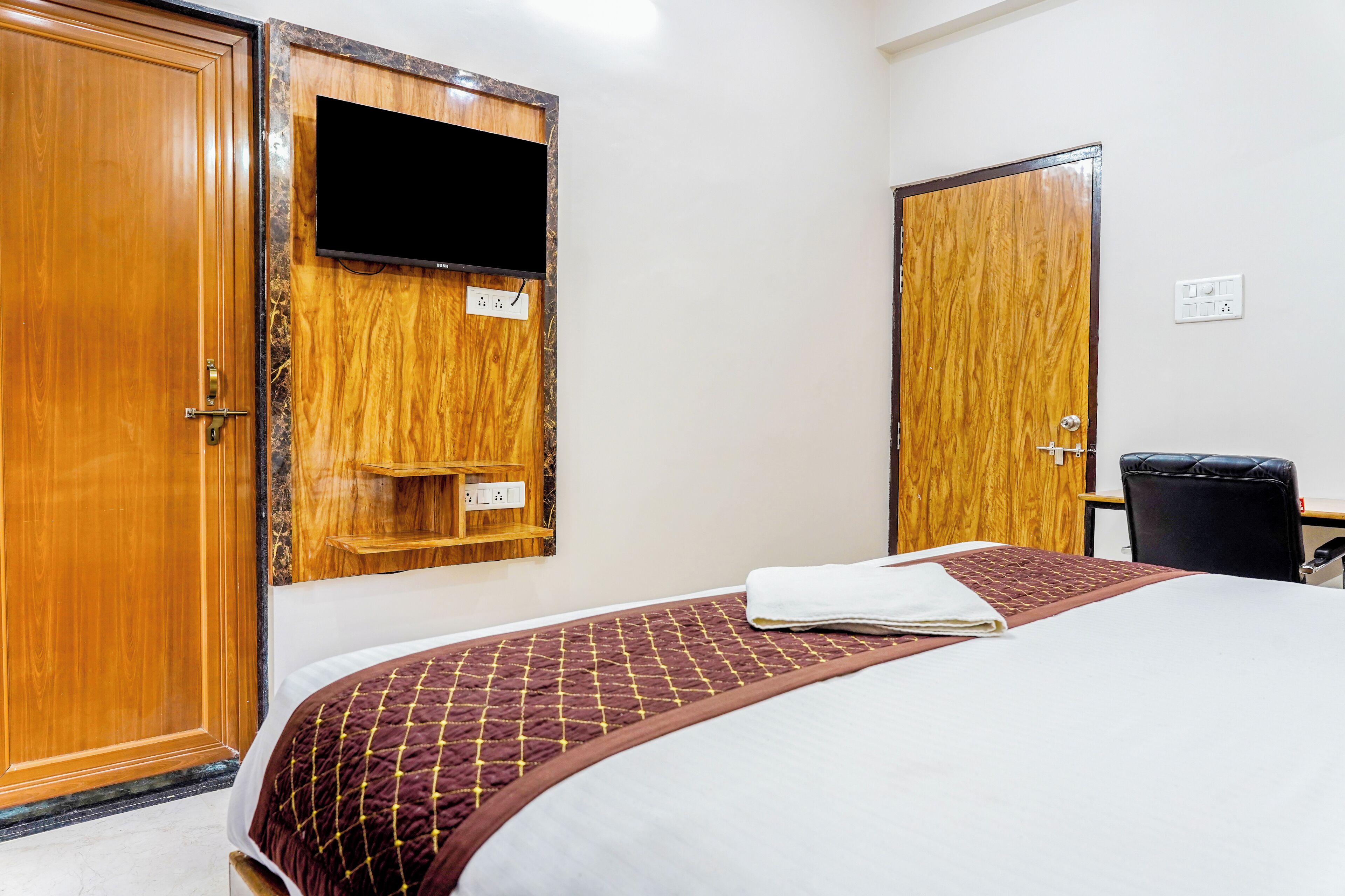 A hotel room with a bed, a TV mounted on a wooden panel, a desk with a chair, and a wardrobe.