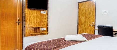 A hotel room with a bed, a TV mounted on a wooden panel, a desk with a chair, and a wardrobe.