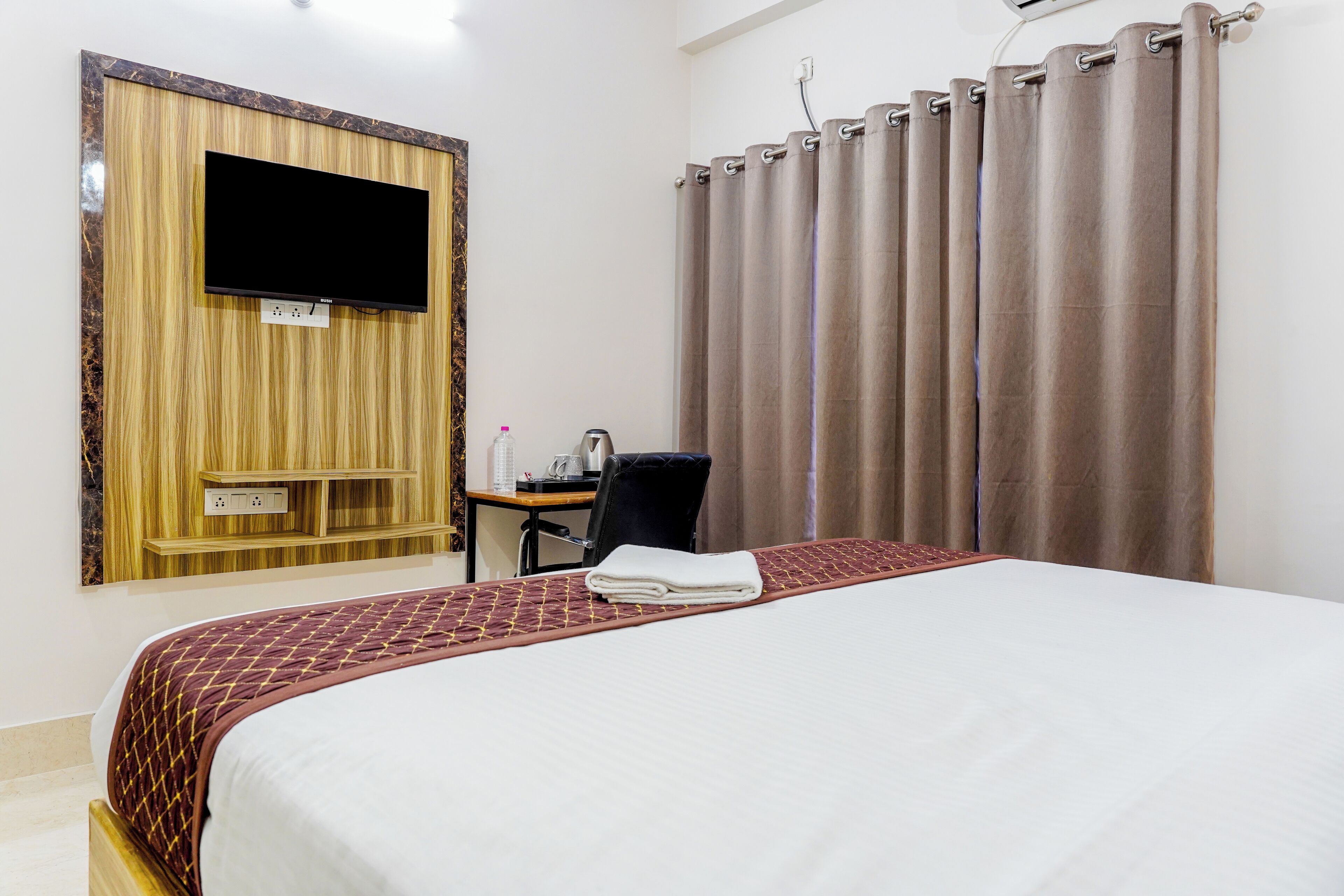 Ein Hotelzimmer mit Bett, Fernseher an einer Wand aus Holzpaneelen, Schreibtisch mit Stuhl und einem Vorhangfenster.