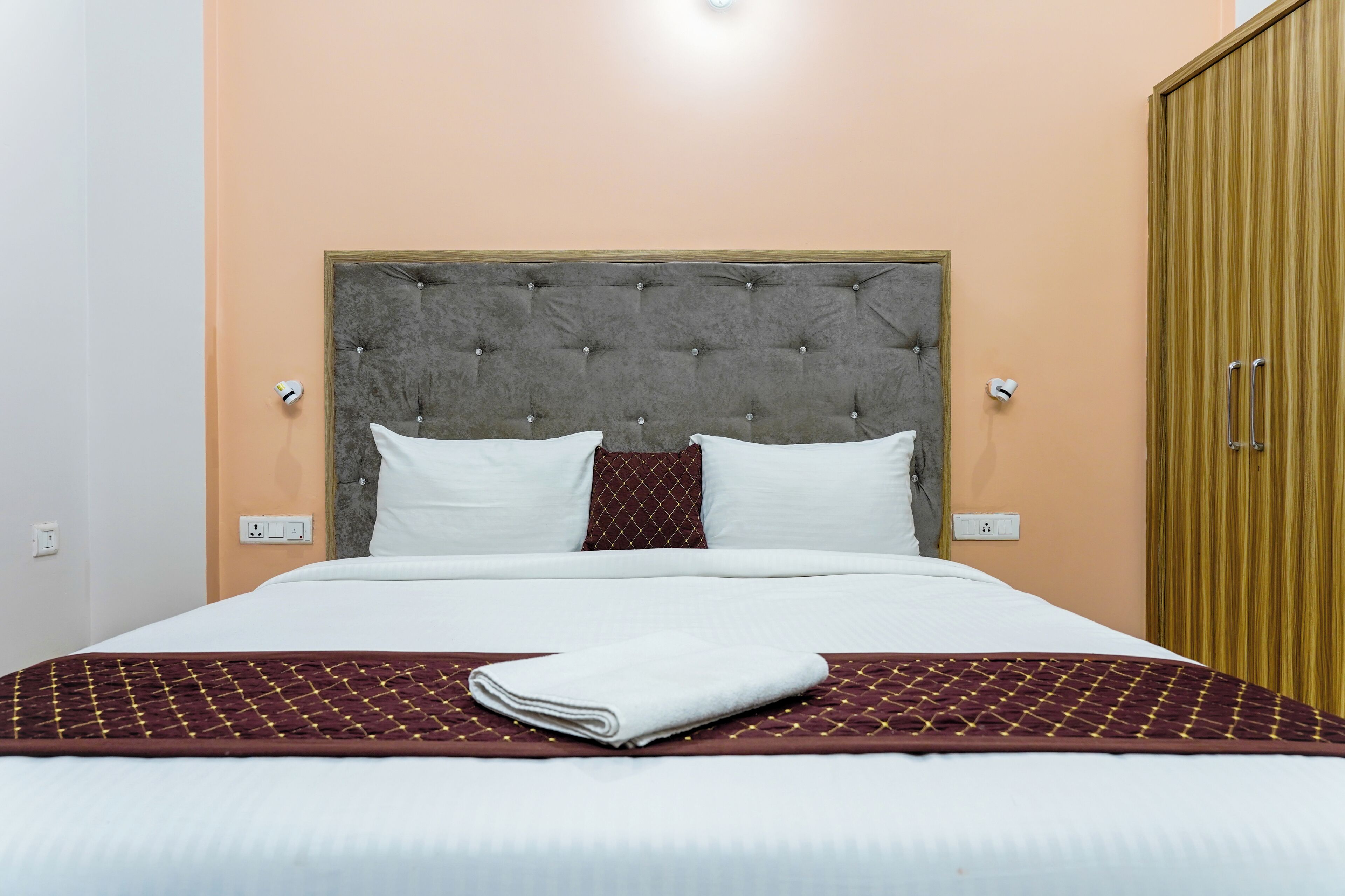 A neatly made bed with a grey tufted headboard, white pillows, and a folded white towel on a patterned bedspread.