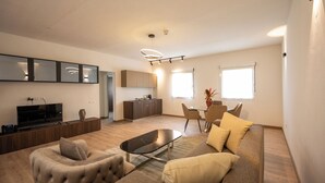 A modern living room with a sofa, armchair, and a coffee table. There is a TV, a dining table with chairs, and a kitchen area with cabinets and a countertop. - Heliconia Park Hotel & Golf Resort (Port Harcourt)