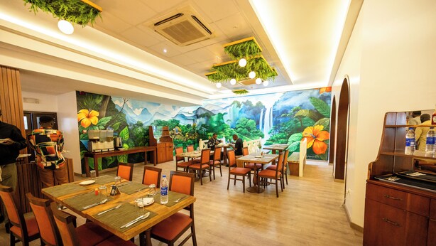 A restaurant with wooden tables and chairs, a mural of a tropical scene, and hanging greenery on the ceiling. - Heliconia Park Hotel & Golf Resort (Port Harcourt)