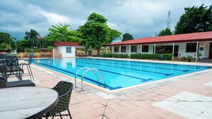 A swimming pool with a tiled deck, outdoor seating, and a building with a red roof. - Heliconia Park Hotel & Golf Resort (Port Harcourt)