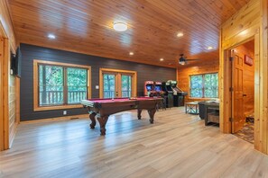 A wooden interior with a pool table, arcade machines, and large windows.