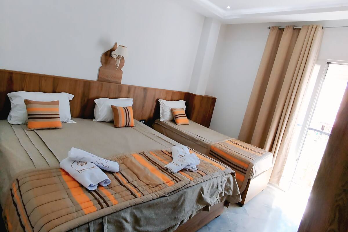 Une chambre d’hôtel avec un grand lit, une tête de lit en bois, un climatiseur fixé au mur et une grande fenêtre avec des rideaux.