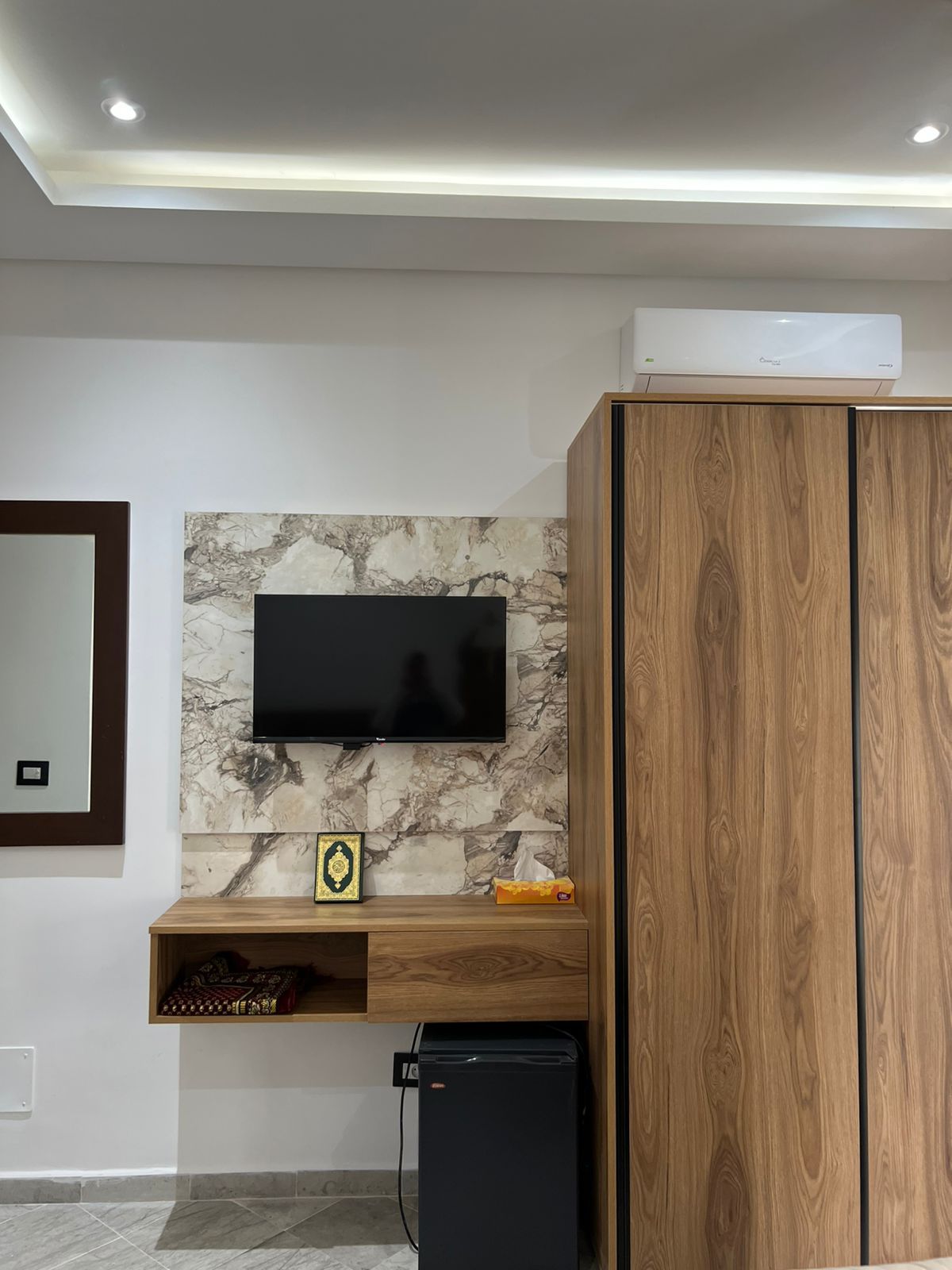 A modern living room with a flat-screen TV mounted on a marble wall, wooden cabinets, a small refrigerator, and a shelf with decorative items.