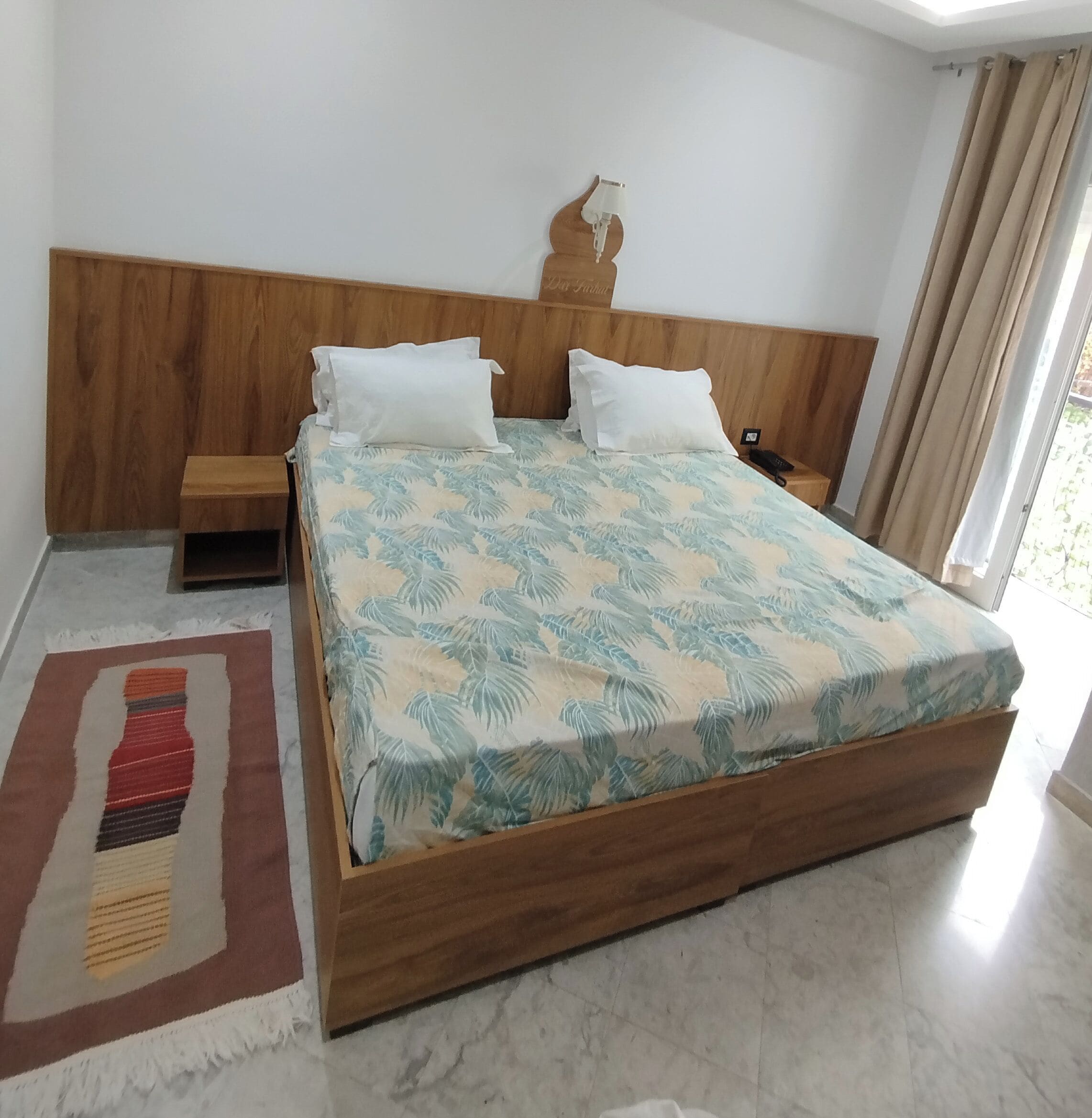 Una cama con colcha estampada con motivos de hojas, cabecera de madera, mesita de noche y velador con lámpara.