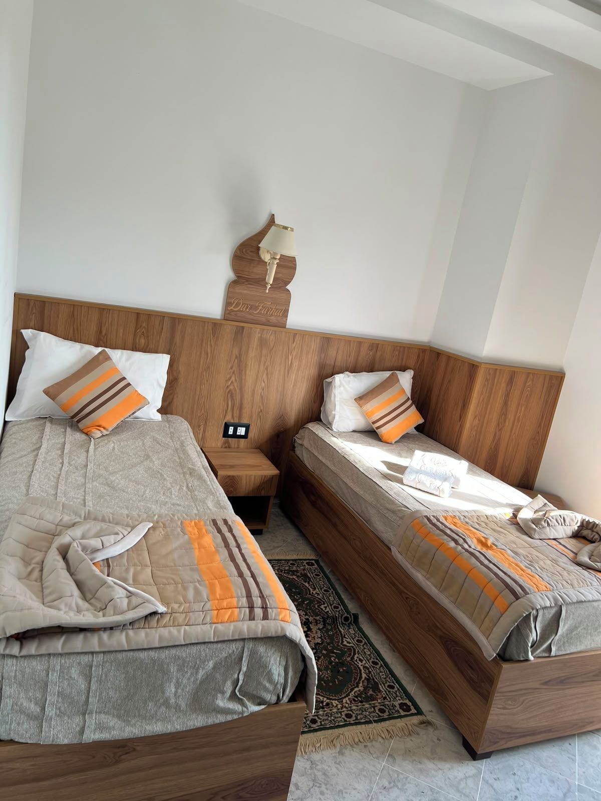 A hotel room with two beds, a wooden headboard, a nightstand, a lamp, and a patterned rug.