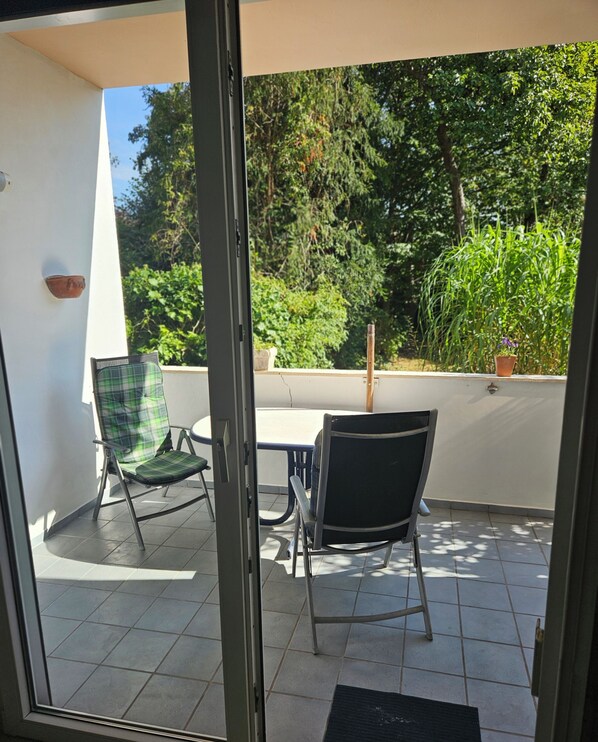 A balcony with two chairs and a table, a door, and a view of greenery.