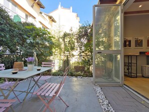 An outdoor patio with a table and chairs, surrounded by greenery and a glass enclosure.