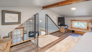 Ein Schlafzimmer im Spitzboden mit schrägen Wänden, Holzbalken, einer Treppe mit Metallgeländern, einem großen Fenster und einem Kamin.