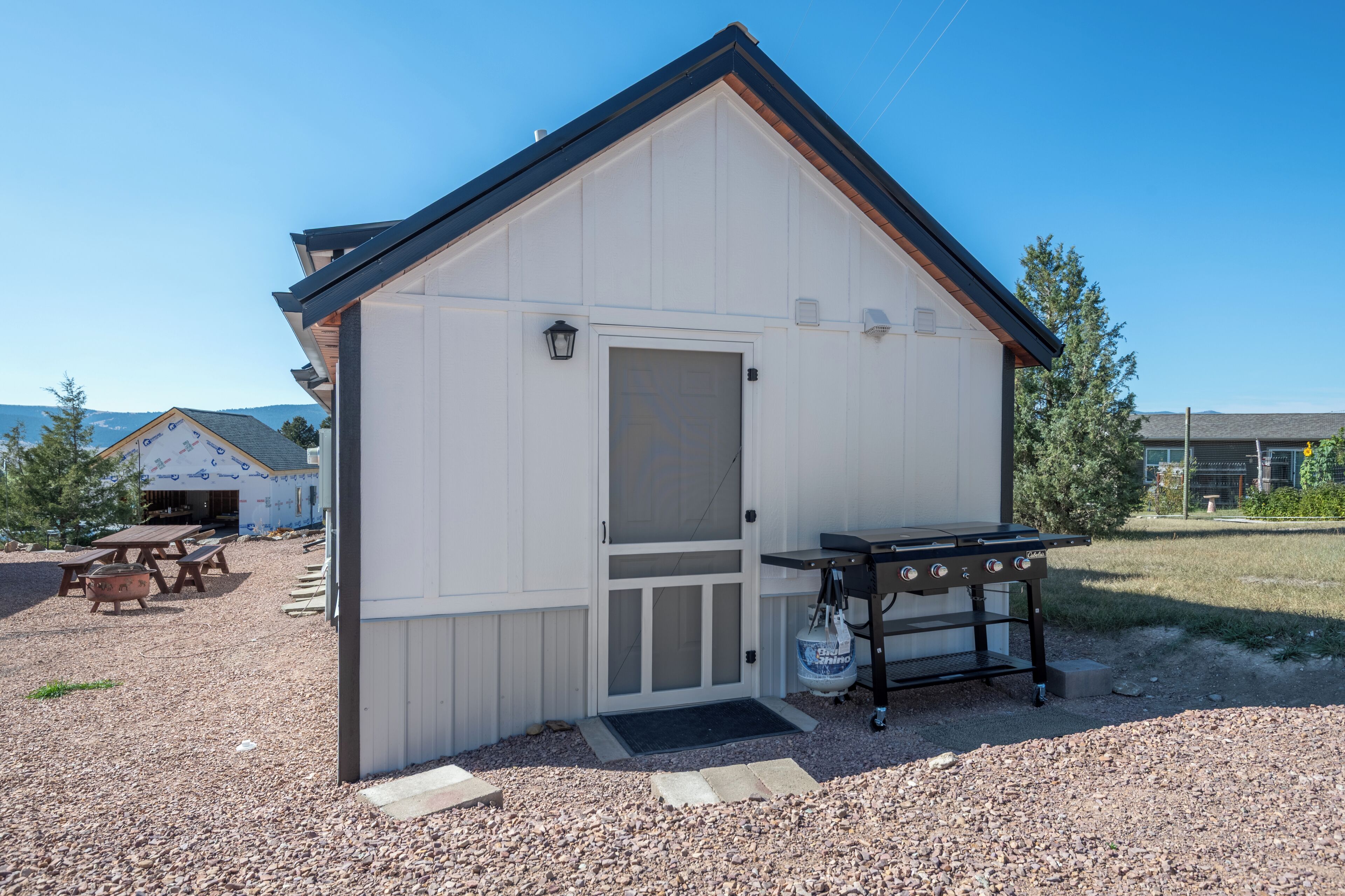 A small, single-story building with a white exterior and a black roof. It features a door with a glass panel, a small porch area, and a barbecue grill outside.