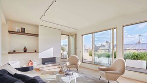 A spacious living room with a sofa, two armchairs, a coffee table, and a fireplace. Large windows offer a view of the cityscape.