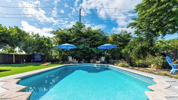 Une piscine rectangulaire dont le bord est recouvert de carreaux, entourée d’une terrasse en pierre. Deux parasols bleus offrent de l’ombre.