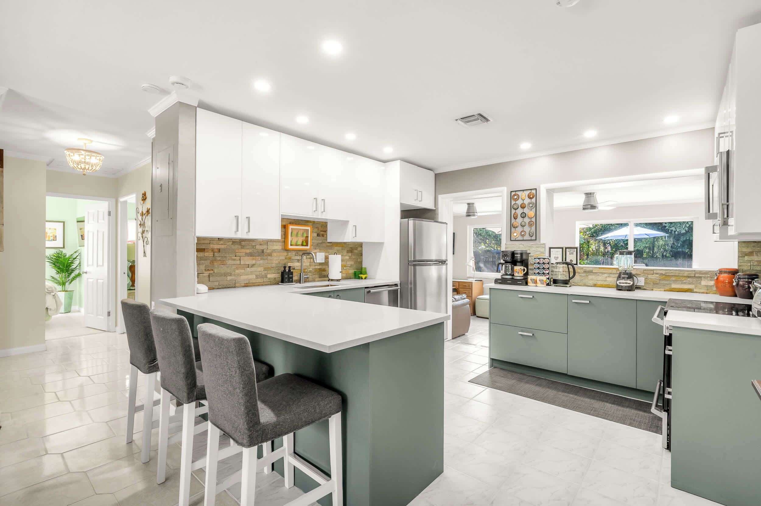 A modern kitchen with white cabinets, a central island with bar stools, a refrigerator, and a window with a view of greenery.